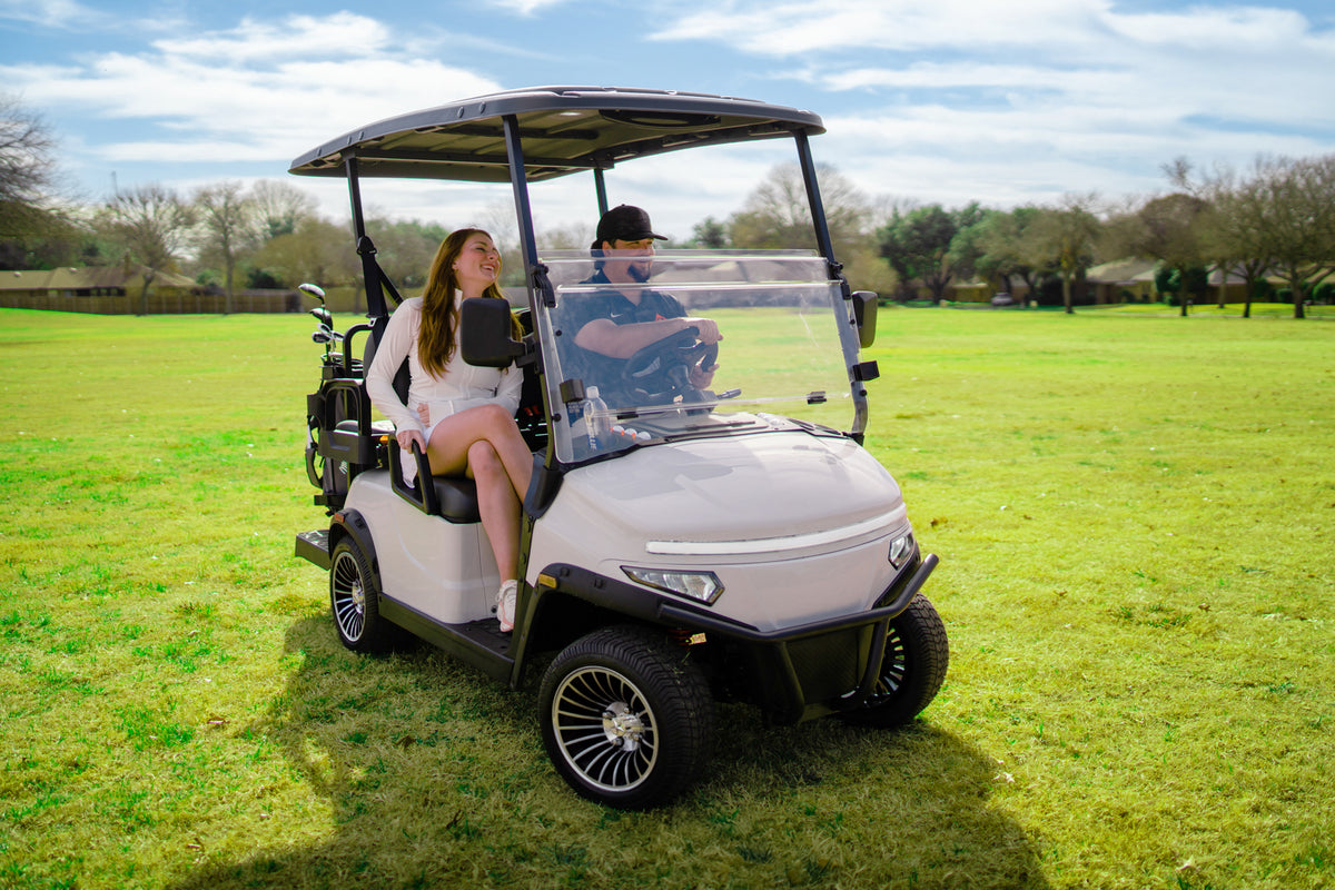 Two people seated in a light-coloured Denago Scout 2 on a sunny golf course